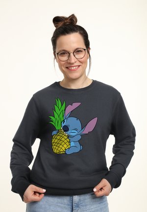 Kvinde med briller og hår i knold, smilende, iført en mørk sweatshirt med en farverig tegneseriefigur, der krammer en ananas.