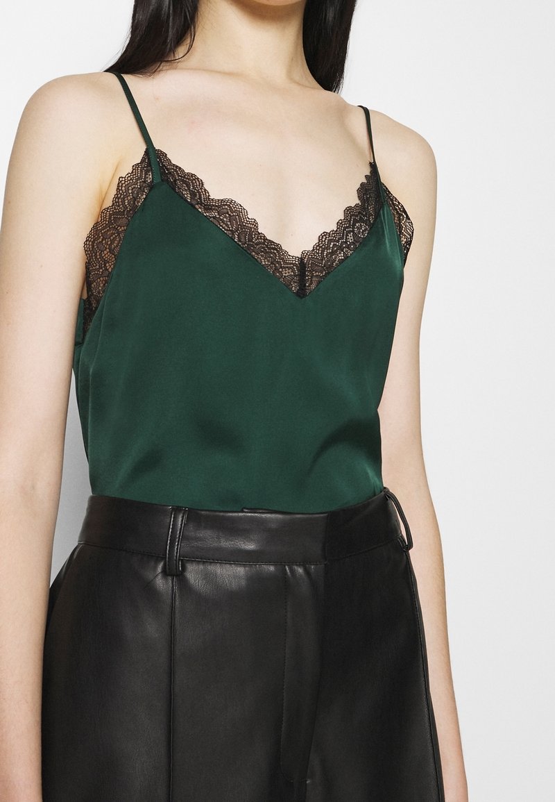 Camisole en satin vert avec un décolleté en V, ornée d'une bordure en dentelle noire le long des bords. Portée avec un pantalon en cuir noir taille haute.