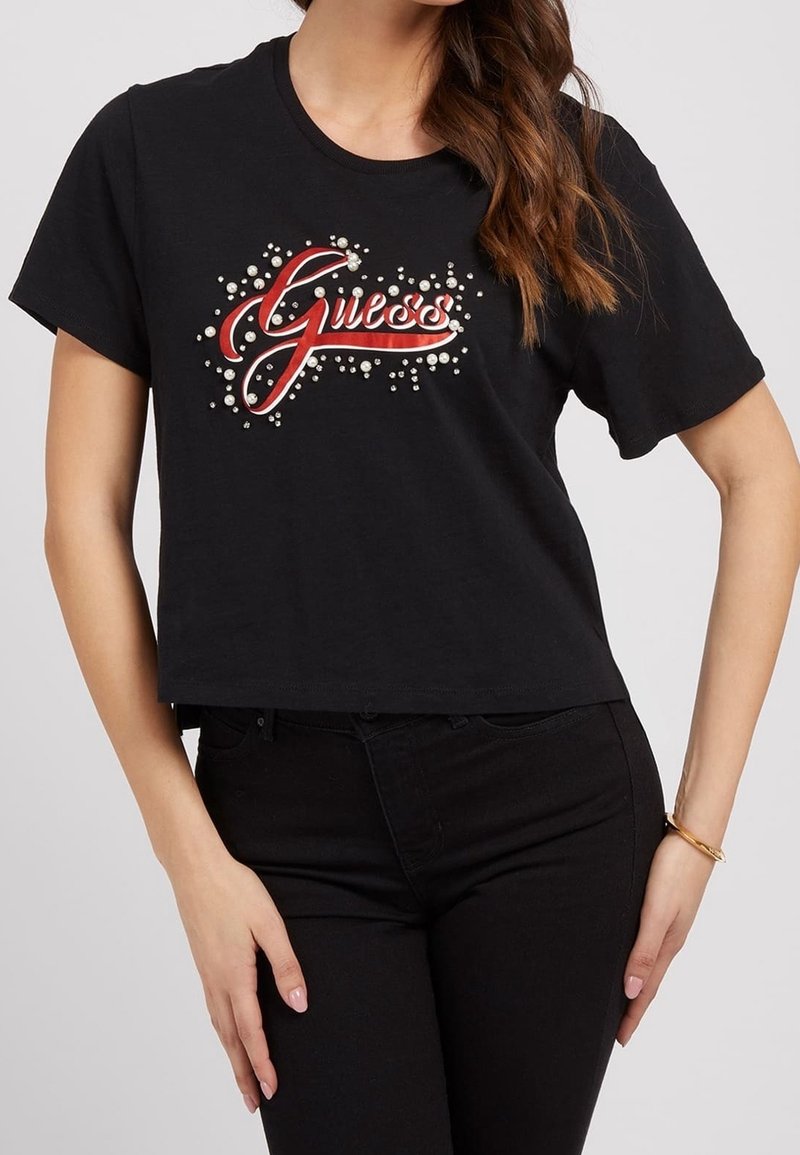 Camiseta negra corta con un logo rojo de "Guess" adornado con cuentas blancas decorativas. Mangas cortas y un escote redondo. Adecuada para el uso casual.