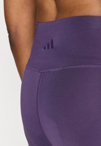 Ljubičaste atletske tajice s visokim strukom, koje imaju teksturiranu tkaninu i Adidas logotip u tamnijoj nijansi na struku.