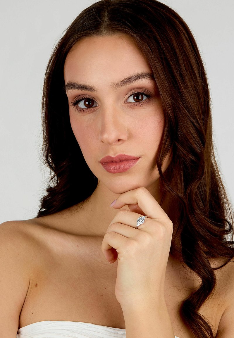 Anello di fidanzamento in argento con una grande pietra centrale, montato su una fascia liscia. Il modello posa con la mano sul mento, mettendo in mostra l'anello.