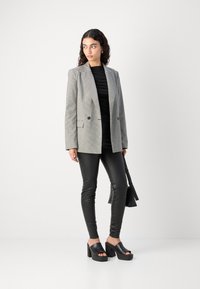 Hall houndstooth mustriga bleiser kaherealise topelt nööpide ja mustade kitsaste retuusidega, kõrgete kontsadega kingadega, kandes musta käekotti.