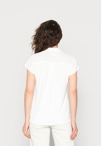 Camicia bianca a maniche corte con un colletto, tessuto liscio e dettaglio sulla schiena. Il design presenta una vestibilità comoda e una trama morbida.