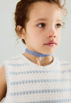 Jeune enfant aux cheveux bouclés portant un haut sans manches blanc à rayures bleu clair et un collier ras du cou bleu avec un petit pendentif en argent.