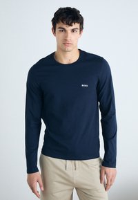 Chemise à manches longues marine en tissu doux, avec un col rond et un petit logo blanc sur la poitrine gauche. Associée à un pantalon beige.