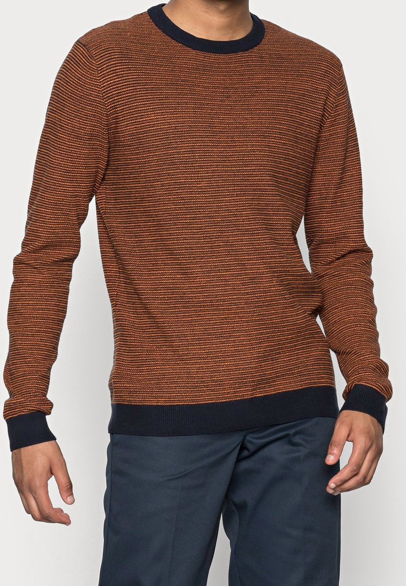 Randig orange och marinblå tröja med rund hals. Stickad struktur och ribbade ärmslut. Buren över mörk marinblå byxor.