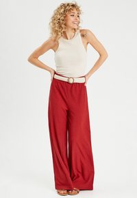 Pantalones rojos de pierna ancha con una tela texturada, combinados con una camiseta sin mangas beige. Un cinturón trenzado blanco con una hebilla redonda acentúa la cintura.