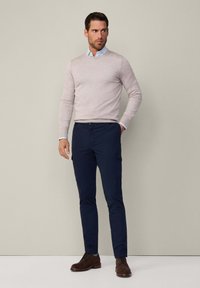 Maglione beige chiaro, camicia blu, pantaloni cargo blu navy e scarpe stringate marroni. L'outfit è completato da uno sfondo neutro.