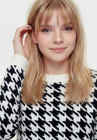 Mladá žena s blond vlasmi v čierno-bielom svetri s psím zubom, jemne si dotýka vlasov a mäkko sa usmieva do kamery.