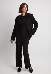 Črn krojen blazer s strukturiranim dizajnom, v kombinaciji s črnimi širokimi hlačami. Obe sta izdelani iz gladkega materiala. Špicaste črne čevlje.