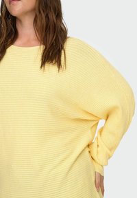 Moteris su rudais plaukais, vilkinti laisva, dryžuota šviesiai geltona megztine, viena ranka remiasi į klubą, stovinti priešais paprastą foną.