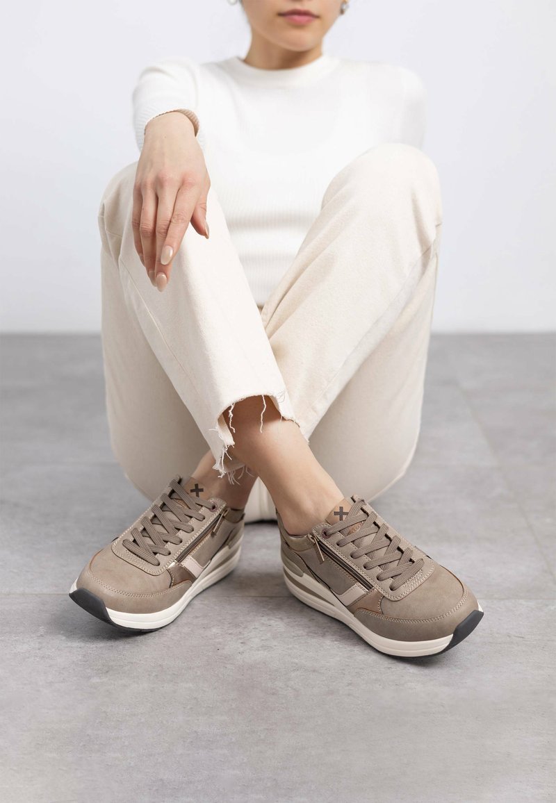 Zapatos beige de cordones con un upper texturizado y acentos contrastantes. Cuentan con una suela acolchada y un sutil detalle de cremallera lateral, colocados sobre un suelo gris.