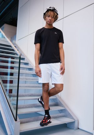Zwart T-shirt, witte shorts en zwart-rode sneakers. Model staat op een trap met een glazen leuning, tegen een lichtgekleurde muur.