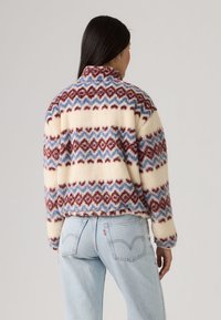 Φλις πουλόβερ με ψηλό γιακά, διαθέτοντας κρεμ βάση, γεωμετρικά κόκκινα και μπλε μοτίβα, και απαλό, υφασμάτινο φινίρισμα.