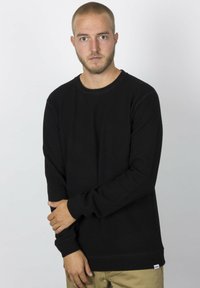 Schwarzes Sweatshirt mit langen Ärmeln und Rundhalsausschnitt, aus dickem Stoff gefertigt. Mit einem kleinen Logostick am Saum. Kombiniert mit beigen Hosen.