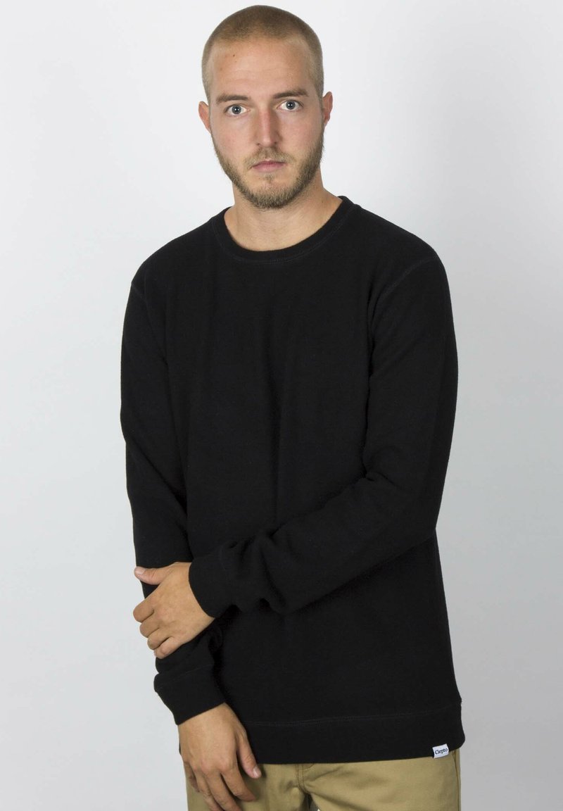 Schwarzes Sweatshirt mit langen Ärmeln und Rundhalsausschnitt, aus dickem Stoff gefertigt. Mit einem kleinen Logostick am Saum. Kombiniert mit beigen Hosen.