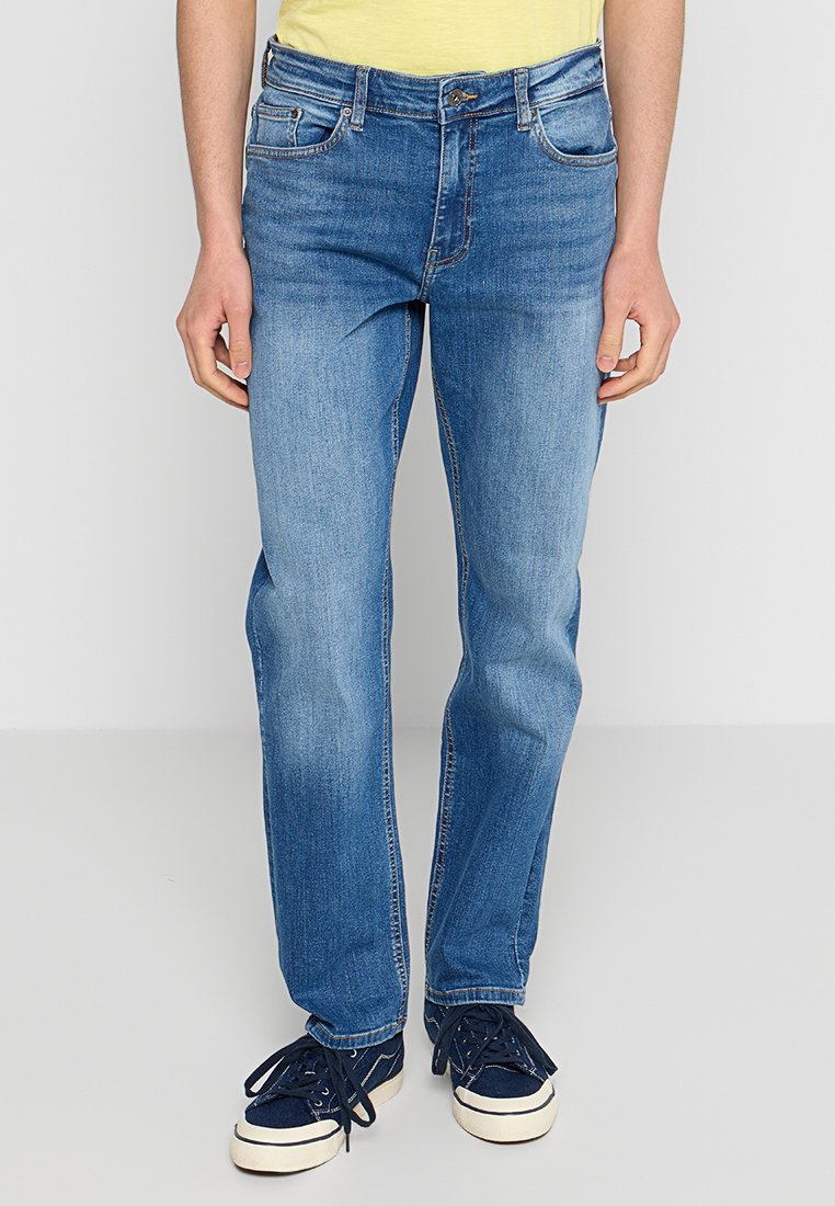 Jeans de mezclilla azul con un corte recto, que presentan bolsillos cosidos, cremallera y botón en la bragueta, y un ligero desgaste. Llevados con zapatillas de deporte azul marino.