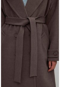Brązowy wełniany płaszcz z szerokim kołnierzem, z paskiem w talii i dwoma przednimi kieszeniami. Posiada guziki na rękawach oraz miękką, fakturowaną tkaninę.