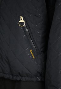 Zwarte gewatteerde jas met een diagonale ritssluiting met gouden hardware en een "Barbour"-logo in geel aan de voorkant.