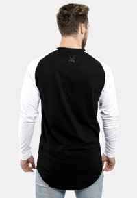 Crna i bijela majica dugih rukava s zaobljenim rubom, s crnim raglan rukavima i malim logom na stražnjem izrezu vrata.
