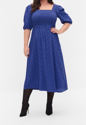 Frau trägt ein blaues Midikleid mit schwarzen Polka-Dots und Puffärmeln, kombiniert mit schwarzen Strumpfhosen und schwarzen spitzen High Heels.