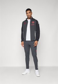 Chaqueta deportiva gris con acentos rojos, logo y rayas blancas, combinada con pantalones grises y zapatillas blancas, con un diseño ajustado.