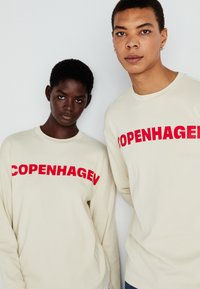 Krém színű, hosszú ujjú pólók, amelyeken merész piros "COPENHAGEN" felirat látható elöl, két közeli helyzetben álló személlyel.