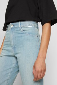 Jeans de ganga azul claro com uma cintura alta, design de cinco bolsos e uma textura ligeiramente desbotada. Um botão metálico e uma etiqueta branca são os detalhes que se destacam.