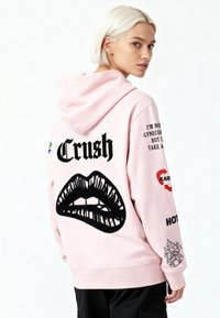 Giovane con capelli corti e biondi che indossa una felpa con cappuccio rosa con grafica di labbra nere e la scritta "Crush" sul retro, che guarda oltre la spalla.