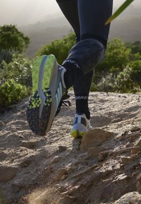 Zapatilla de trail running con parte superior gris, detalles en negro y una suela texturizada que presenta elementos de agarre en amarillo y negro, mostrada en movimiento sobre terreno rocoso.