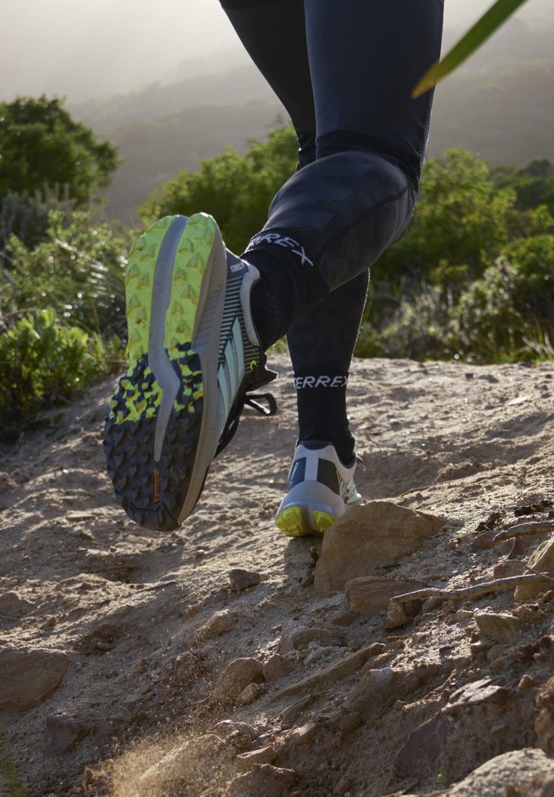 Zapatilla de trail running con parte superior gris, detalles en negro y una suela texturizada que presenta elementos de agarre en amarillo y negro, mostrada en movimiento sobre terreno rocoso.