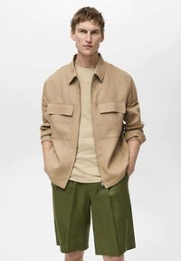 Beige lynlåsjakke med to forlommer, båret over en lys tan skjorte, parret med grønne shorts, der viser en afslappet og casual pasform.