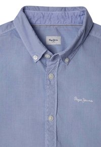 Šviesiai mėlynas marškinis su apykakle, pagamintas iš sklandžios medžiagos. Apatinės dalies balti mygtukai ir siuvinėtas „Pepe Jeans“ logo.