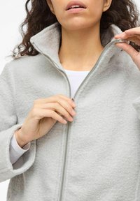 Gaiši pelēka fliša jaka ar augstu apkakli un rāvējslēdzēja aizdari, ar teksturētu virsmu un brīvu piegriezumu. Rokas, kas rāda rāvējslēdzēja regulēšanu.