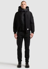 Czarna kurtka puchowa z kapturem nałożona na pikowaną kamizelkę. Czarne dżinsy i czarne botki dopełniają outfit. Prosty design z funkcjonalnymi zamkami.