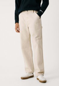 Beige Baumwoll-Twill-Hosen im gerade geschnittenen Stil, mit Seitentaschen und einem dezenten Etikett am Bund; kombiniert mit weißen Sneakern.