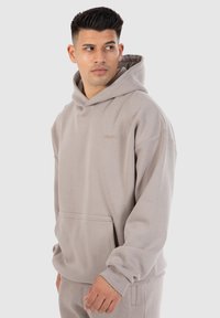 Hellgrauer Kapuzenpullover mit einer vorderen Tasche, aus weichem Stoff gefertigt. Hat ein schlichtes Design und gerippte Bündchen. Dezenter gestickter Text auf der Brust.
