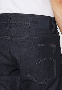 Dark blue denim jeans with a leather waistband patch featuring the embossed logo "RAW" and contrasting stitching on back pockets.