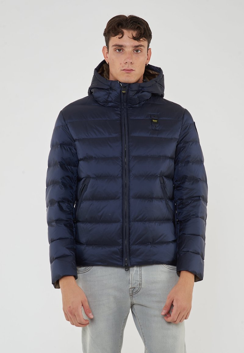 Giovane uomo che indossa un giubbotto imbottito blu navy con cappuccio e jeans grigi chiari, in piedi contro uno sfondo bianco semplice.
