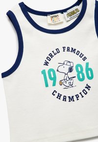 Vit linne med marinblå kant, prydd med Snoopy-design och texten "VÄRLDENS BERÖMDA MÄSTARE 1986" i turkos och svart. Tillverkad av bomull.