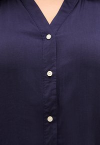 Κοντινό πλάνο ενός μπλε πουκάμισου με V-λαιμόκοψη και τρία λευκά κουμπιά κατά μήκος της μπροστινής πλευράς.