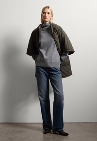 Giacca trapuntata di un verde oliva scuro, maglione grigio con orlo a costine, jeans blu e scarpe scure. Tessuti lisci, vestibilità rilassata e colori neutri.