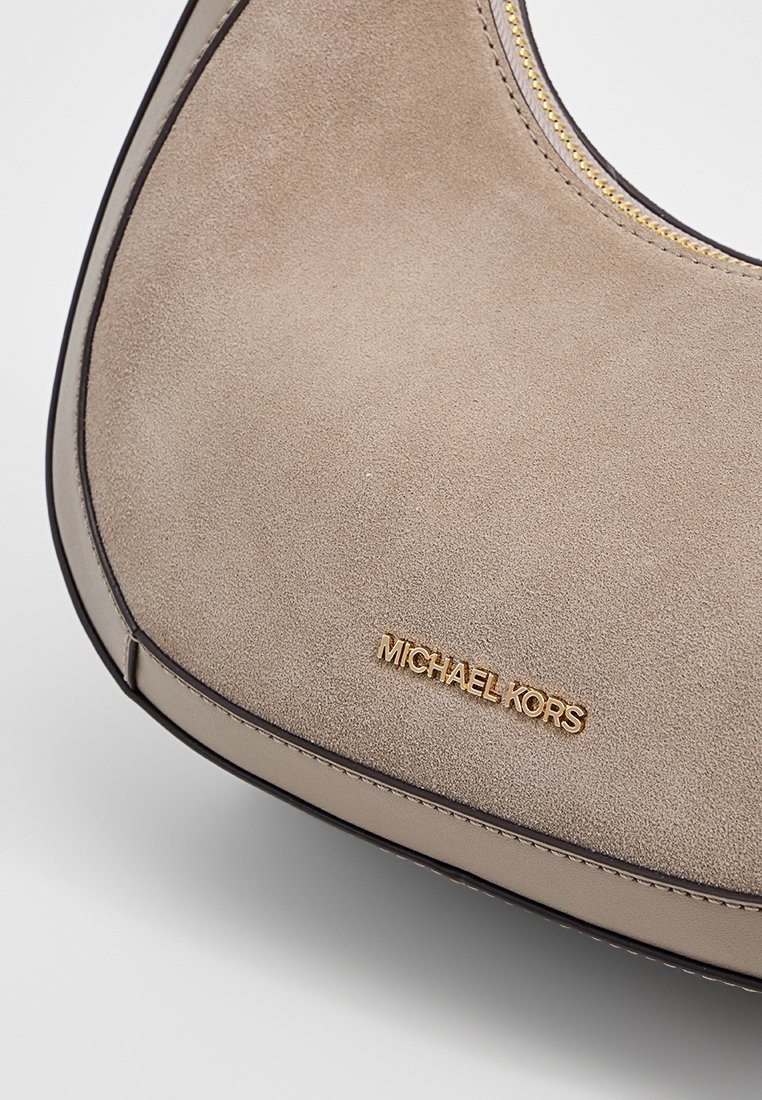 Beige suède schoudertas met een gebogen vorm, voorzien van een goudkleurig logo en ritsen, accentueert door donkere randen en een soepele textuur.