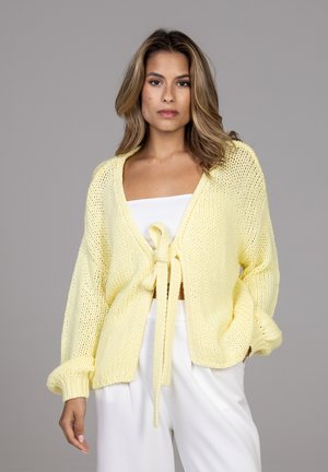 Femme portant un cardigan tricoté jaune clair noué devant par-dessus un haut blanc et un pantalon blanc ample, debout devant un fond gris.