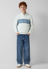 Felpa bianca con cappuccio caratterizzata da una grafica blu "SLVR", abbinata a jeans in denim blu a gamba larga e sneakers bianche con suole color sabbia.