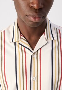 Chemise à manches courtes en tissu écru avec des rayures verticales en rouge, olive, jaune et marine. Col design avec deux boutons à l'avant.