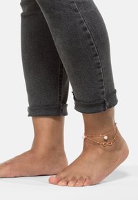 Una cavigliera con doppie catene in oro rosa, adornata con perle e un simbolo dell'infinito, indossata con jeans grigi scuri arrotolati e piede nudo.