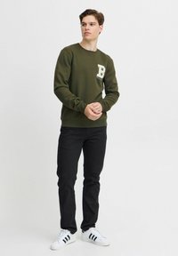 Olivengrøn sweatshirt med en tekstureret 'B'-applikation på brystet, parret med sorte jeans og hvide sneakers med sorte striber.
