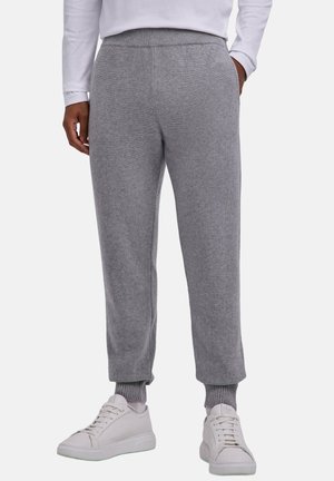 Persoon draagt grijze gebreide broek met geribde boorden en witte sneakers, staat met handen in de zakken.