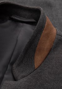 Color de chaqueta gris oscuro con un forro negro suave y un acento de ante marrón. Presenta un tejido texturizado y un diseño estructurado.